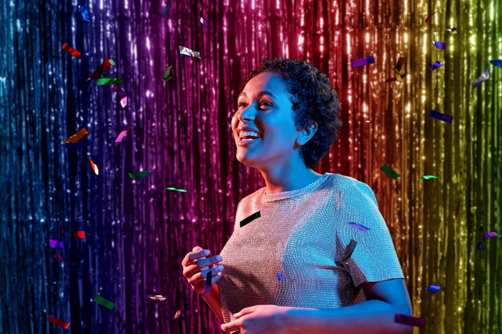 leisure, party and nightlife concept - happy smiling young african american woman under confetti over ultraviolet neon lights over rainbow foil fringe curtain background. african woman under confetti at nightclub party