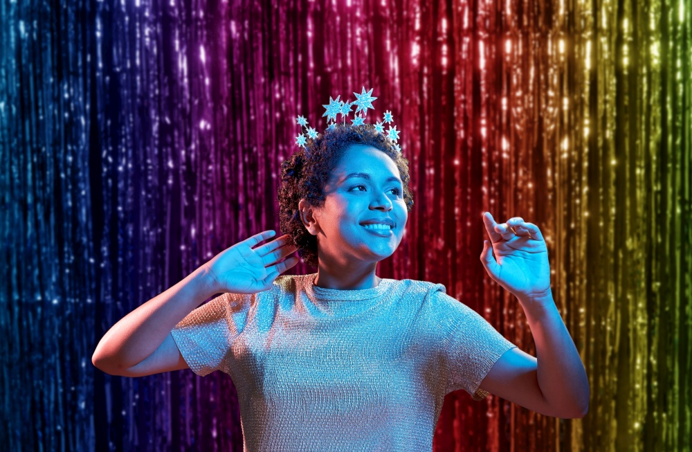 celebration and nightlife concept - smiling young african american woman dancing at christmas or new year party in ultraviolet neon lights over rainbow foil curtain background. african woman at christmas or new year party