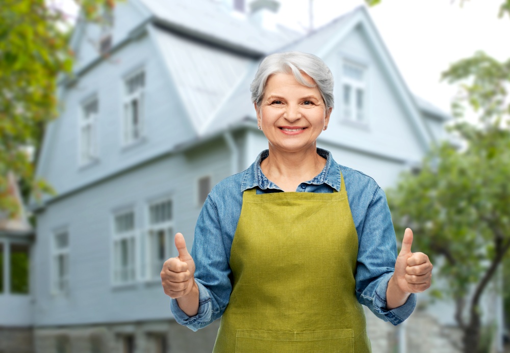 gardening, farming and old people concept - portrait of smiling senior woman in green garden apron showing thumbs up over house background. senior woman in garden apron showing thumbs up