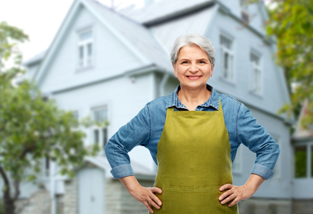 gardening, farming and old people concept - portrait of smiling senior woman in green garden apron over house background. portrait of smiling senior woman in garden apron