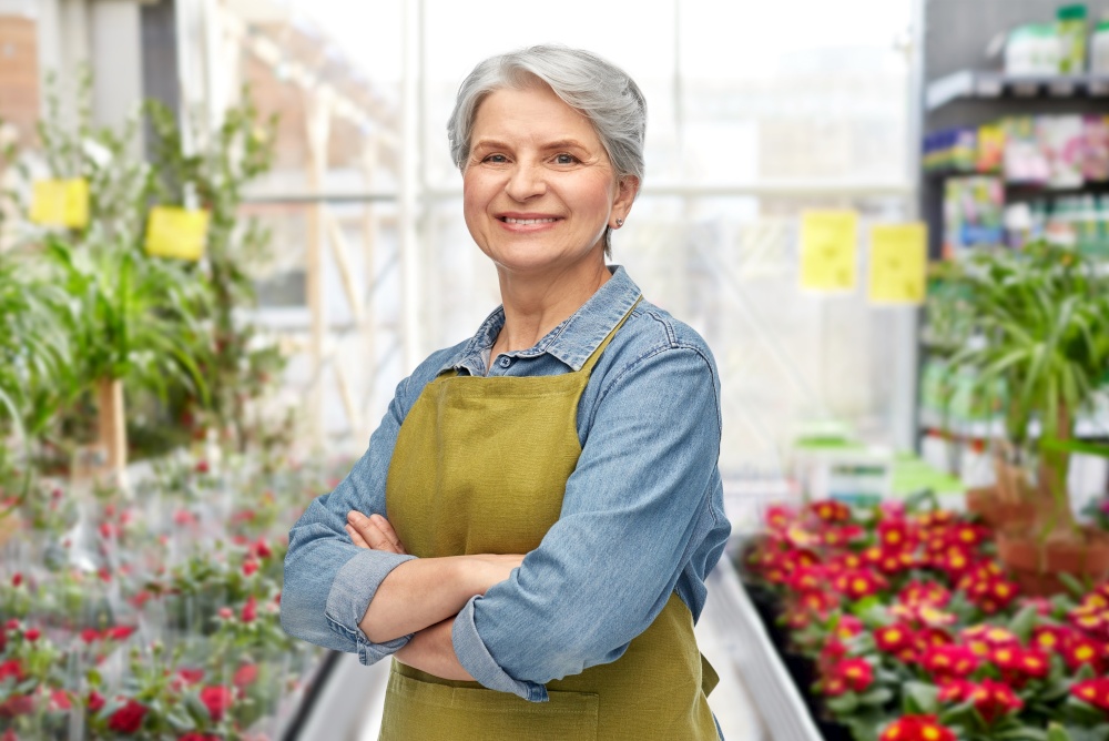 gardening, farming and old people concept - portrait of smiling senior woman in green garden with apron crossed arms over flower shop background. smiling senior woman in gardening center