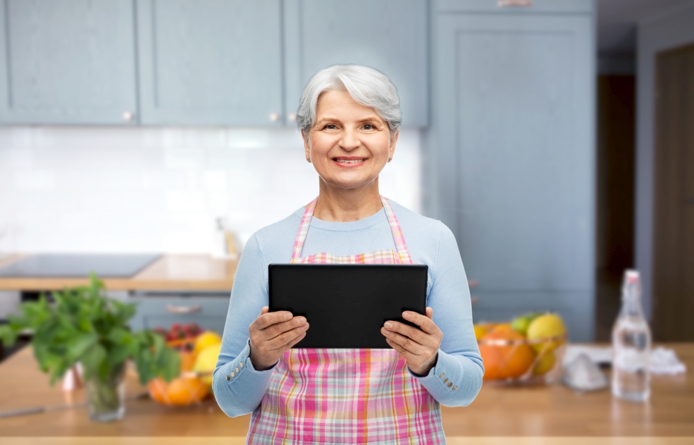 cooking, culinary and old people concept - portrait of smiling senior woman in apron with tablet pc compute over kitchen background. smiling senior woman with tablet pc at kitchen