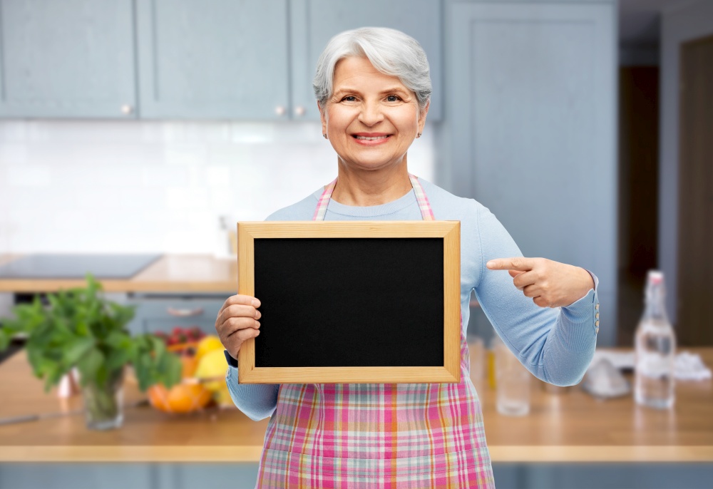 cooking, culinary and old people concept - portrait of smiling senior woman in apron showing chalkboard over kitchen background. smiling senior woman in apron with chalkboard