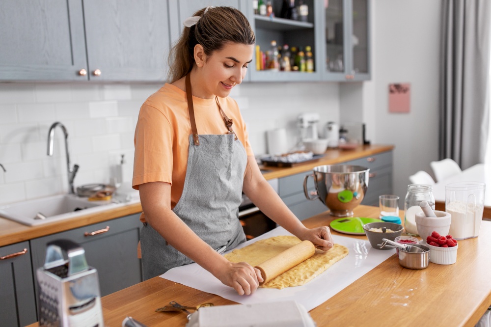 culinary, bake and people concept - happy smiling young woman cooking food on kitchen at home rolls dough with rolling pin. woman cooking food and baking on kitchen at home