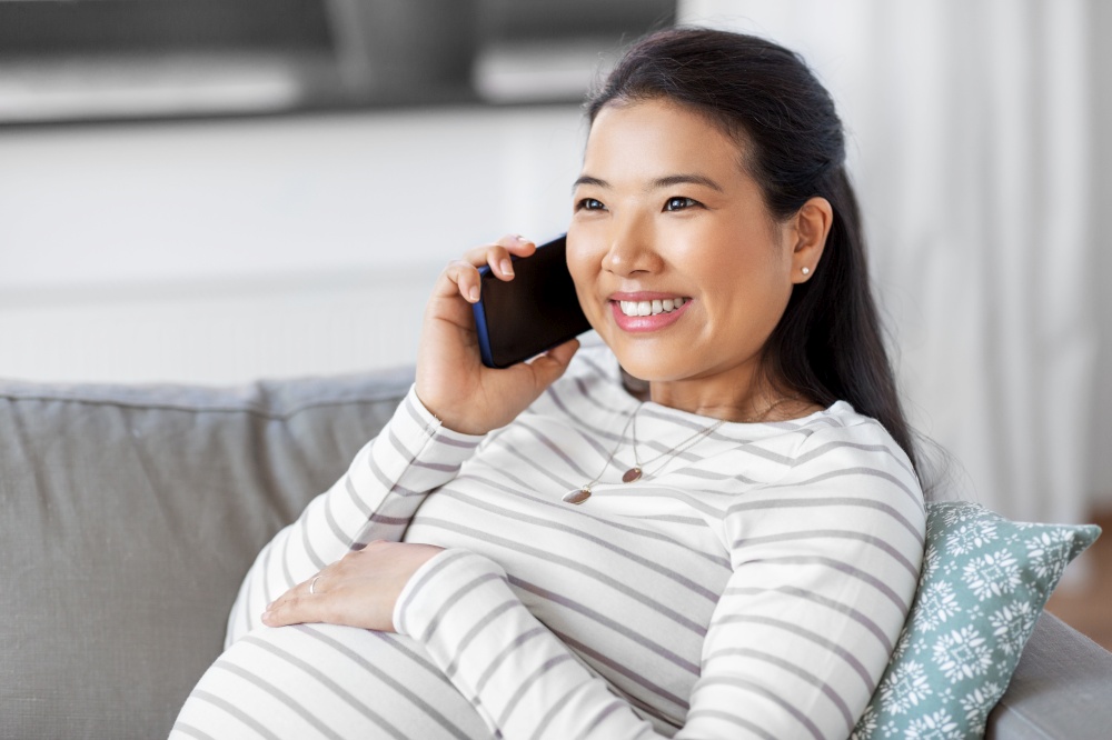 pregnancy, rest, people and expectation concept - happy smiling pregnant asian woman sitting on sofa at home and calling on smartphone. happy pregnant woman calling on smartphone at home