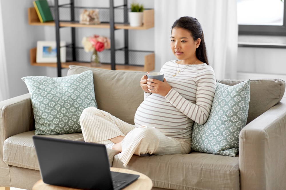 pregnancy, rest, people and expectation concept - pregnant asian woman with laptop computer sitting on sofa at home and drinking tea. pregnant asian woman with laptop at home