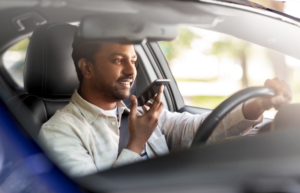 transport, people and technology concept - smiling indian man or driver driving car and recording message using voice command recorder on smartphone. man driving car and recording voice by smartphone