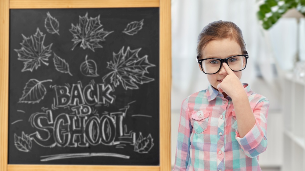 education, learning and people concept - happy little student girl in eyeglasses over chalkboard with back to school lettering on background. little student girl in glasses over chalkboard