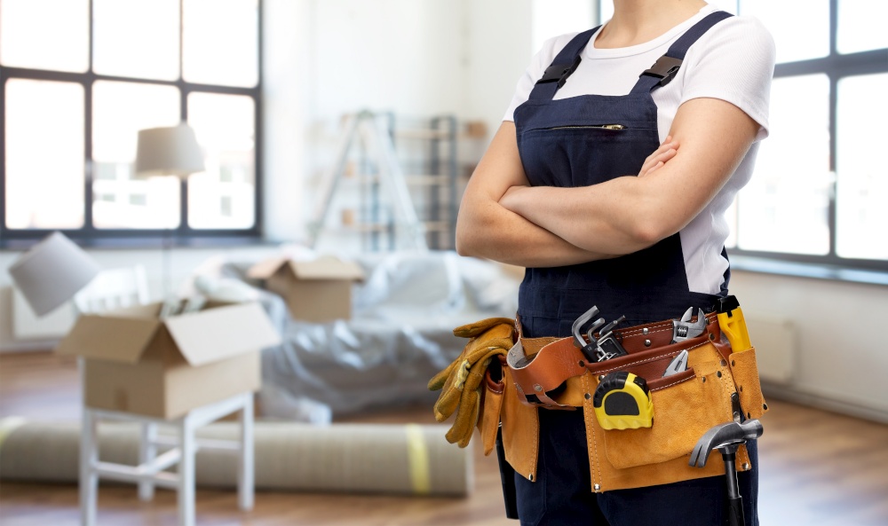 moving, repair and people concept - woman or builder with working tools on belt over new home background. close up of female worker in overall at new home