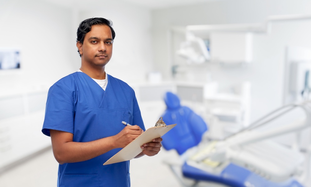 healthcare, stomatology and medicine concept - indian doctor or male dentist in blue uniform writing medical report on clipboard over dental clinic office background. male doctor with clipboard at dental office
