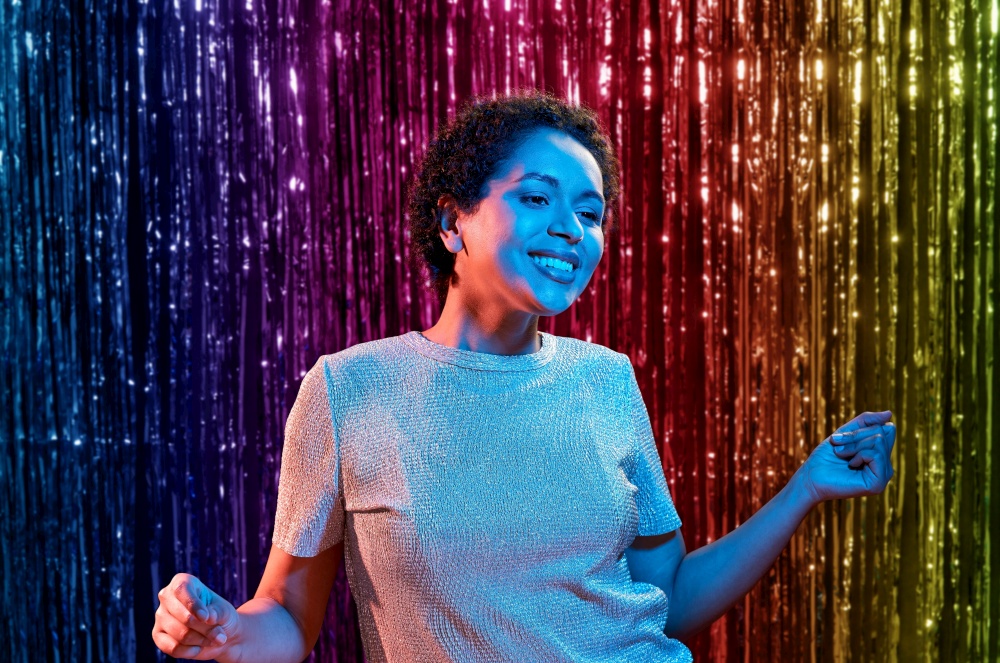 leisure, clubbing and nightlife concept - smiling young african american woman dancing in ultraviolet neon lights at party over rainbow foil curtain background. african american woman dancing at party