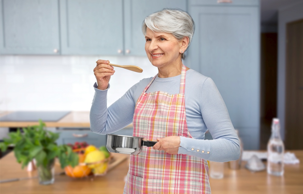 food cooking, culinary and old people concept - portrait of smiling senior woman in apron with pot and spoon over kitchen background. senior woman in apron with pot cooking food