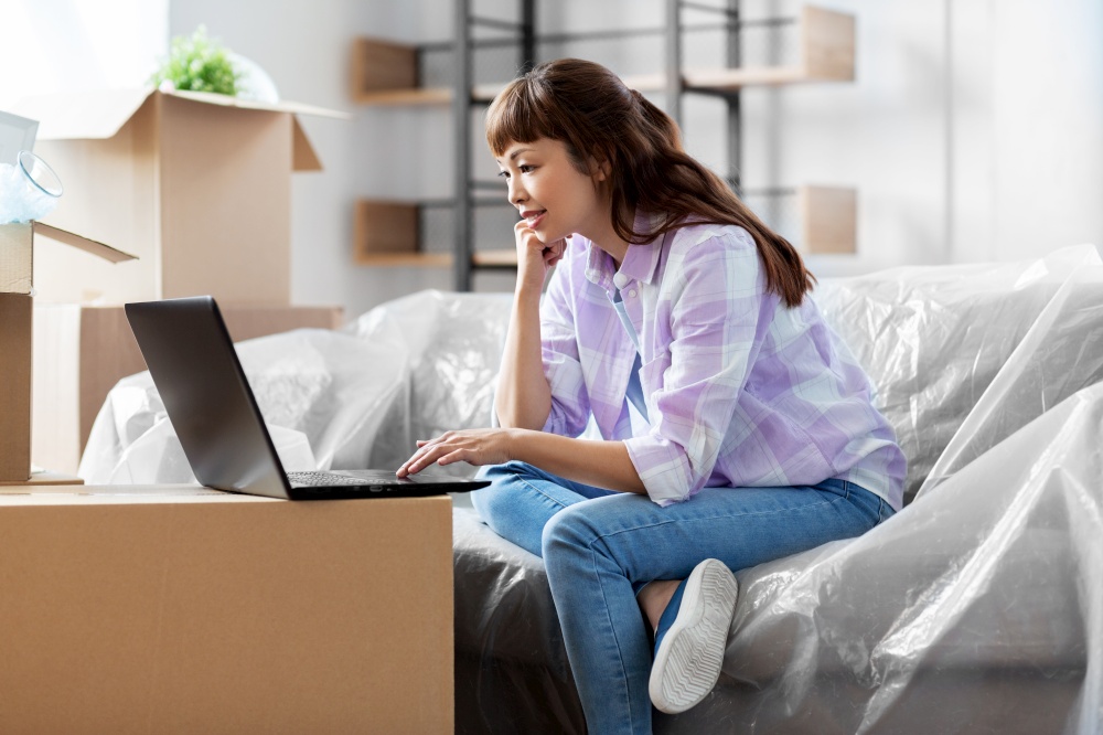 moving, people and real estate concept - happy smiling asian woman with laptop computer and boxes at new home. woman with laptop moving into new home