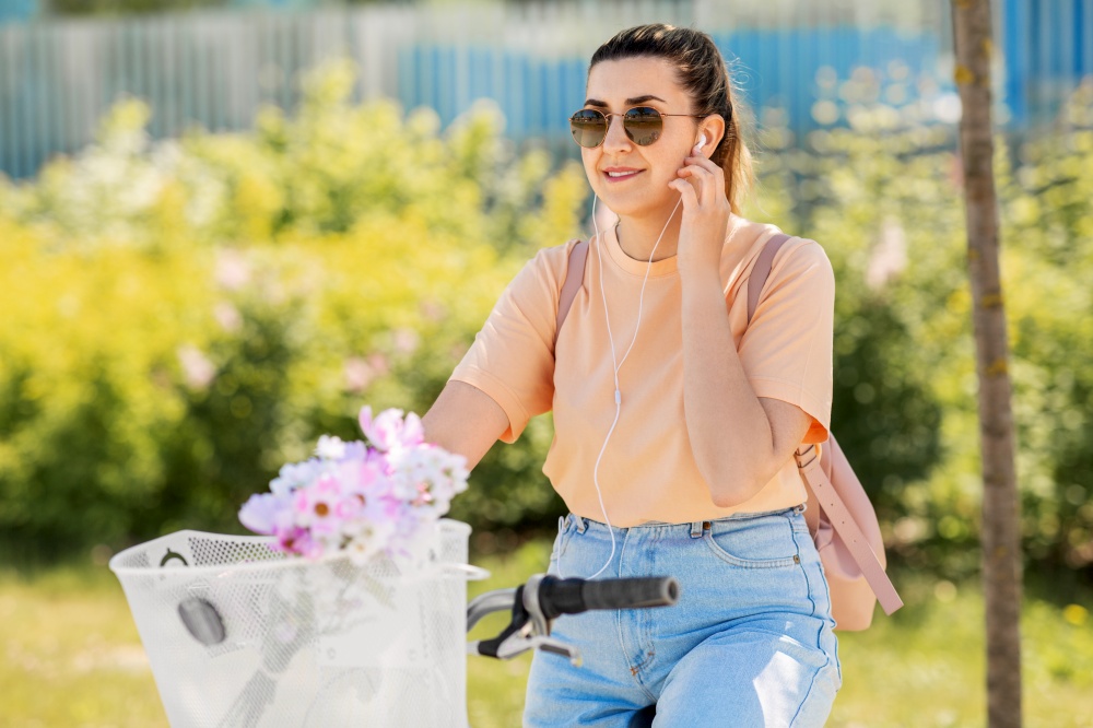 people, leisure and lifestyle - happy smiling young woman with earphones, backpack and flowers in basket riding bicycle on city street. happy woman with earphones riding bicycle in city