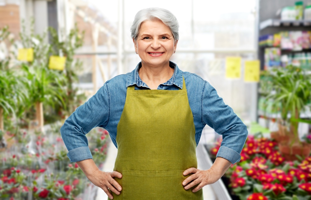 gardening, farming and old people concept - portrait of smiling senior woman in green garden apron over flower shop background. smiling senior woman in gardening center