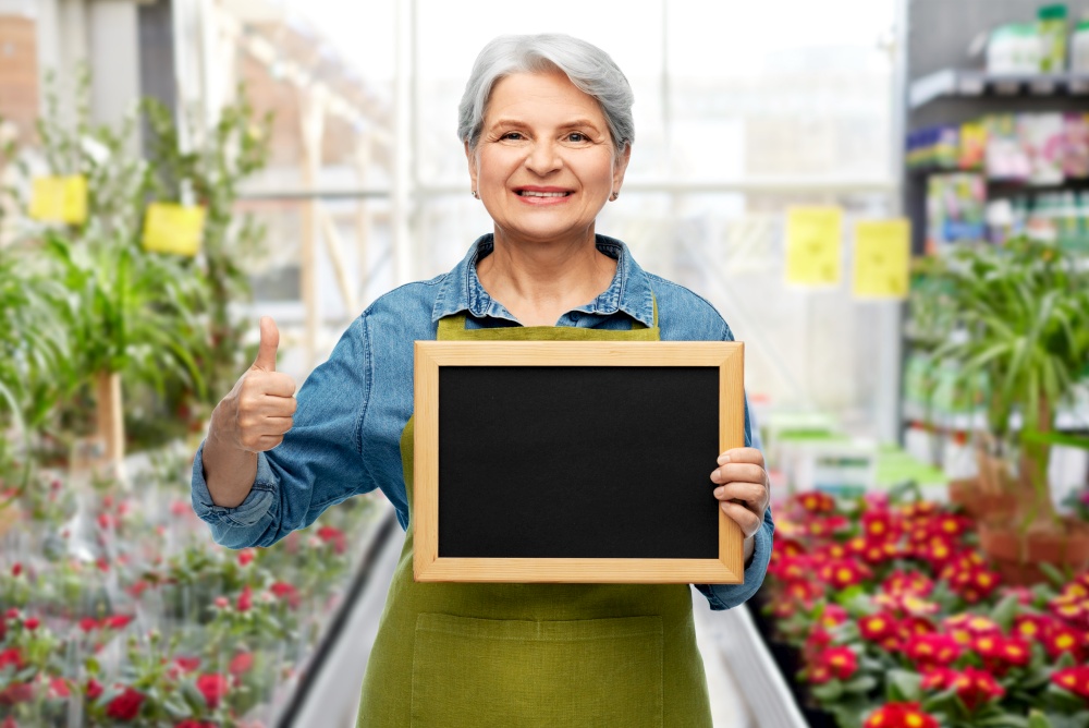 gardening, farming and old people concept - portrait of smiling senior woman in green garden apron holding chalkboard and showing thumbs up over flower shop background. senior gardener with chalkboard showing thumbs up