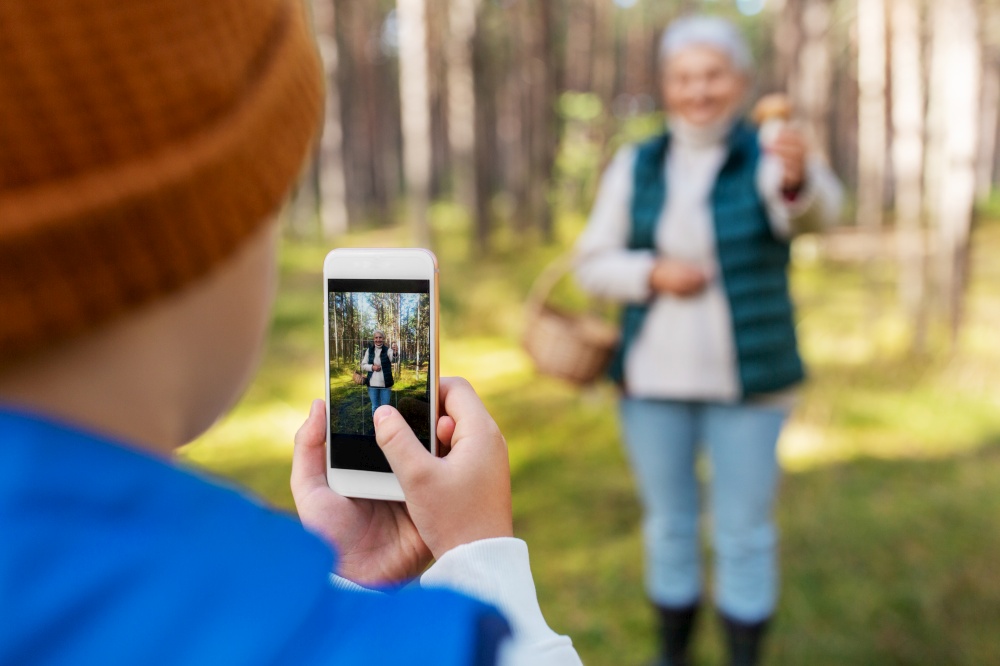picking season, leisure and people concept - grandson with smartphone photographing happy smiling grandmother with mushroom and basket in forest. grandson photographing grandmother with mushroom