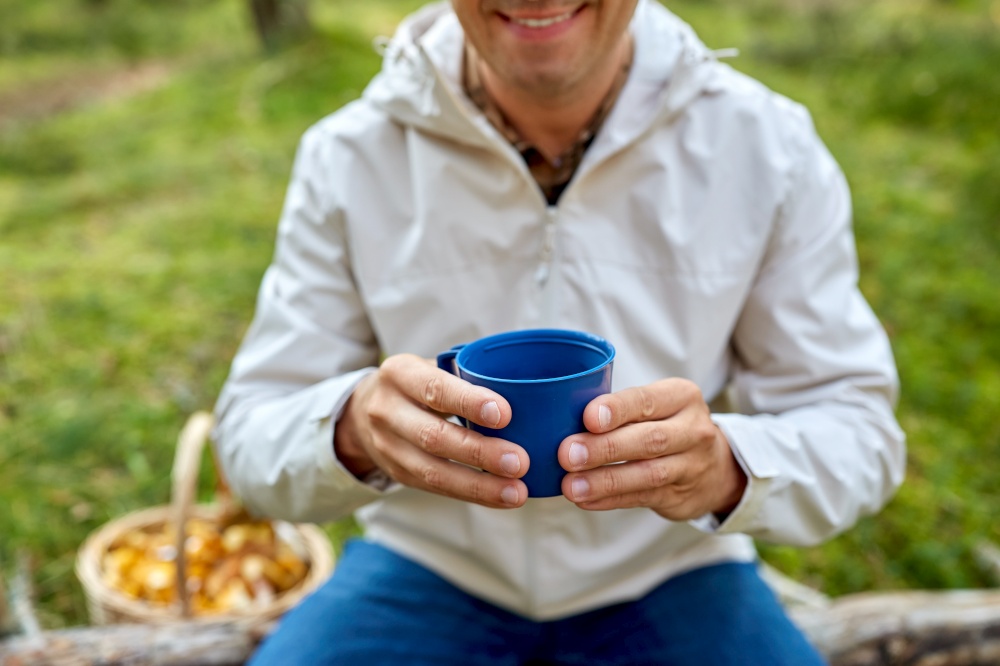 picking season and leisure people concept - close up of man with wicker basket of mushrooms sitting on fallen tree and drinking tea in autumn forest. man with basket of mushrooms drinks tea in forest