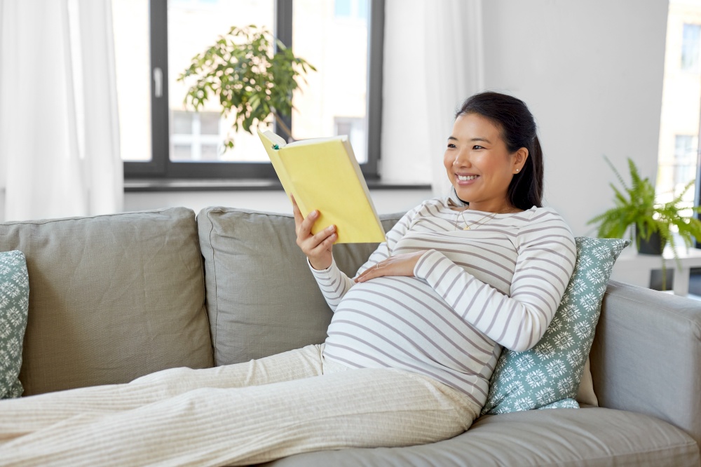 pregnancy, people and leisure concept - happy smiling pregnant asian woman sitting on sofa and reading book at home. happy pregnant woman reading book at home