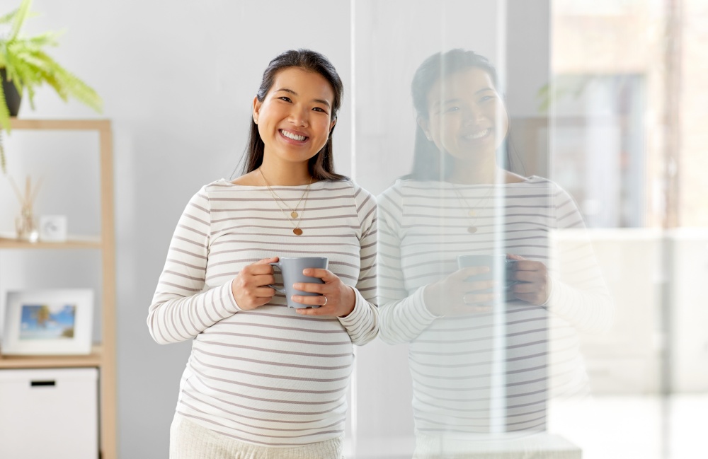 pregnancy, rest, people and expectation concept - happy smiling pregnant asian woman with mug drinking tea at home. happy pregnant woman drinking tea at home