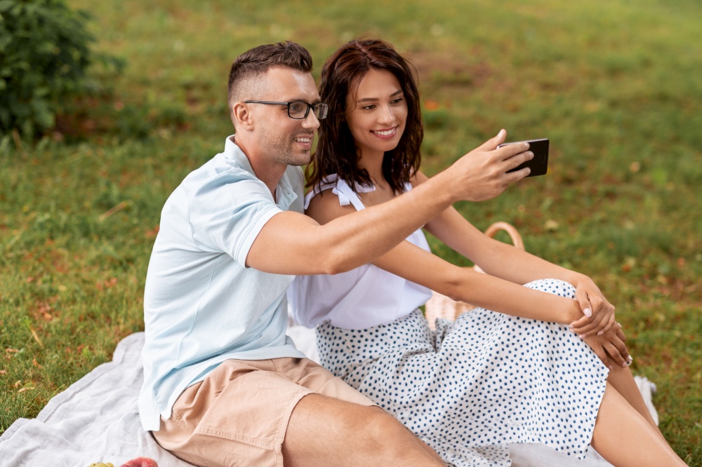 leisure and people concept - happy couple having picnic and taking selfie with smartphone at summer park. happy couple taking selfie at picnic in park