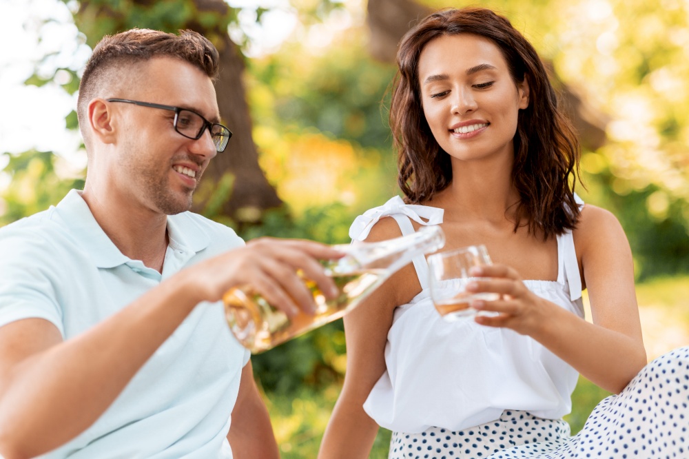 leisure and people concept - happy couple having picnic and pouring wine to glass at summer park. happy couple with wine having picnic at park