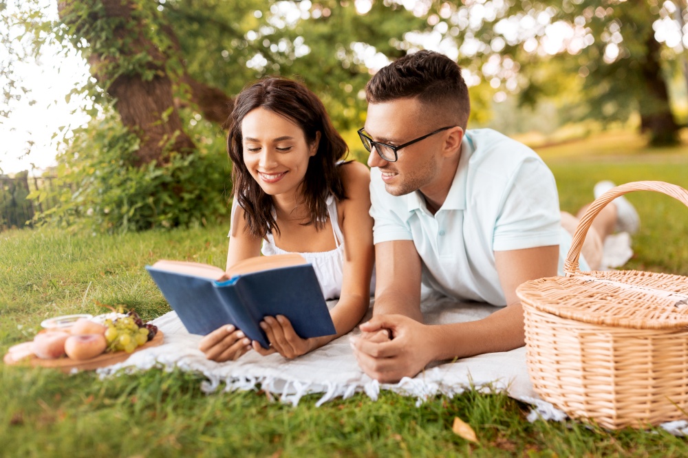 leisure, vacation and people concept - happy couple lying on picnic blanket at summer park and reading book together. happy couple reading book on picnic at summer park