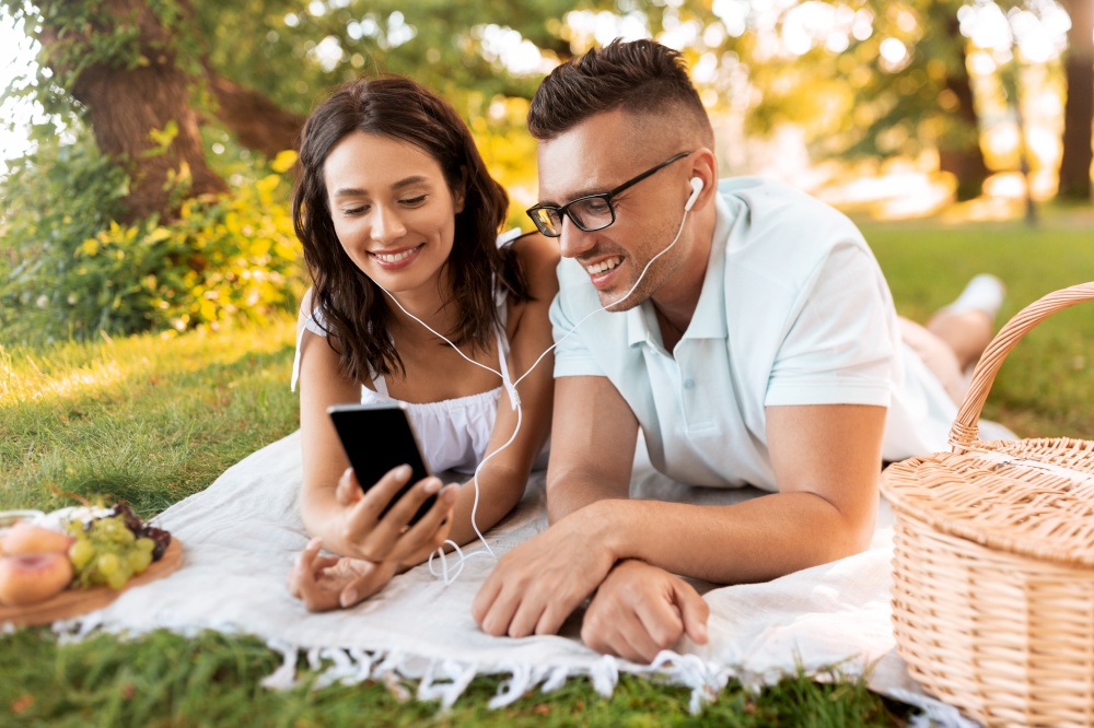 leisure, vacation and people concept - happy couple with earphones and smartphone listening to music on picnic blanket at summer park. couple with earphones and smartphone at picnic