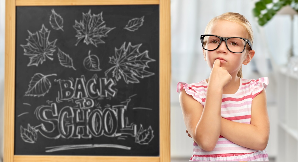 education, learning and people concept - cute little girl in black glasses over chalkboard with back to school lettering on background. little student girl in glasses thinking