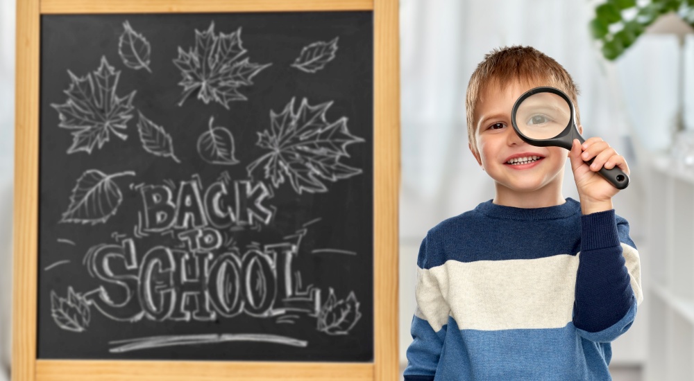 education, learning and people concept - smiling little boy looking through magnifying glass over chalkboard with back to school lettering on background. little student boy looking through magnifier