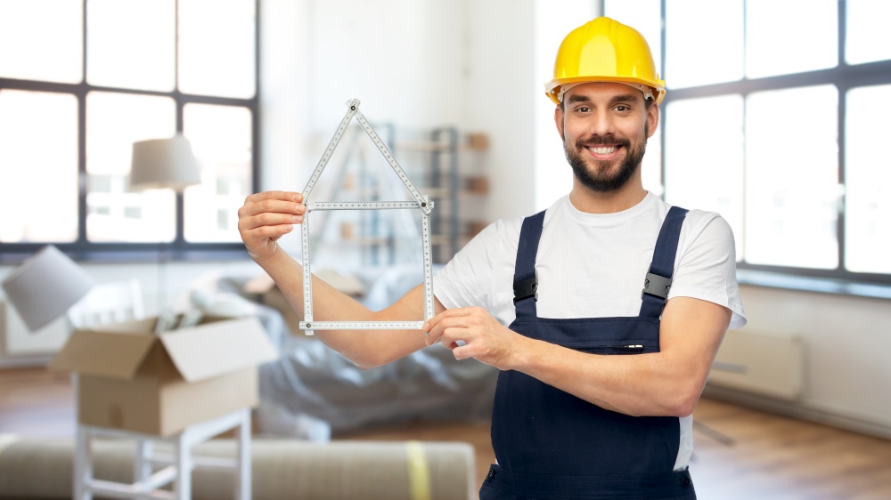 repair, construction and building concept - happy smiling male worker or builder in yellow helmet and overall with carpenter&rsquo;s rule in shape of house over home room background. happy male builder with carpenter&rsquo;s rule at home