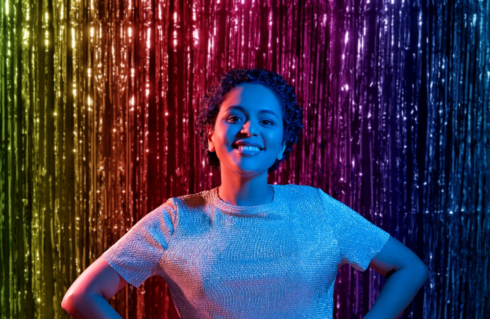 leisure, clubbing and nightlife concept - smiling young african american woman at party in ultraviolet neon lights over rainbow foil curtain background. african woman at party over rainbow curtain