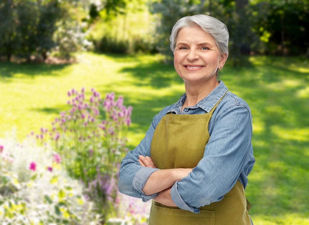 gardening, farming and old people concept - portrait of smiling senior woman in green with apron crossed arms over summer garden background. portrait of smiling senior woman in garden apron