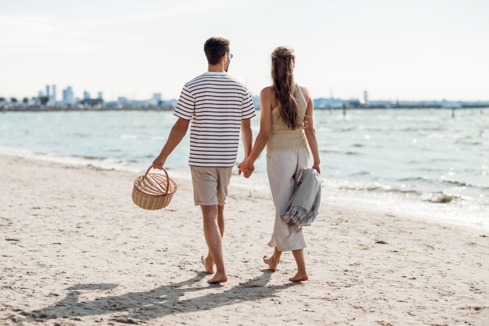 leisure, relationships and people concept - happy couple with picnic basket and blanket walking along beach in tallinn, estonia. happy couple with picnic basket walking on beach