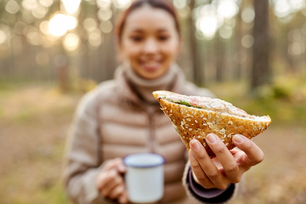 picking season, leisure and people concept - close up of young asian woman drinking tea and eating sandwich in autumn forest. woman drinking tea with sandwich in autumn forest