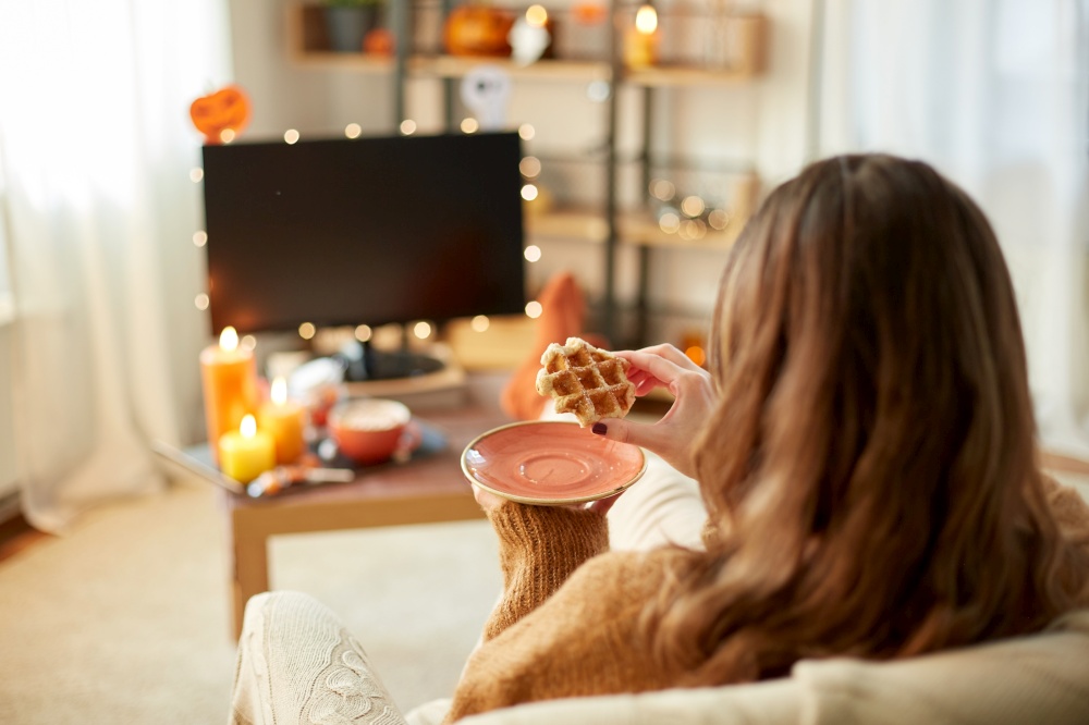 halloween, holidays and leisure concept - young woman watching tv and eating waffle at cozy home. woman watching tv and eating waffle on halloween