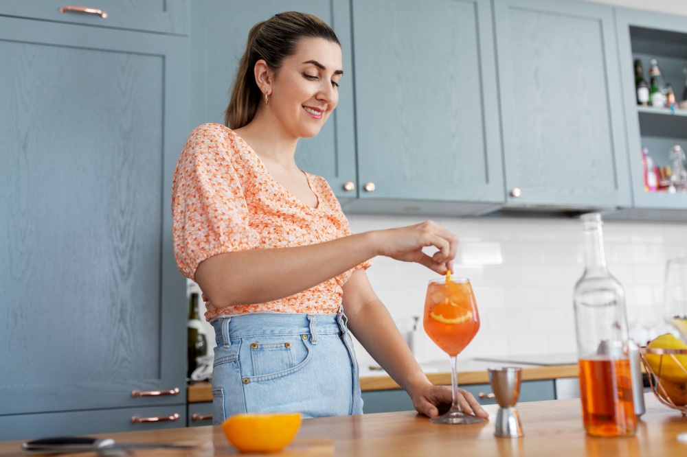 culinary, drinks and people concept - happy smiling young woman making orange cocktail at home kitchen. woman making cocktail drinks at home kitchen