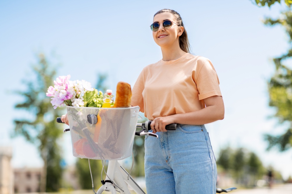 people, leisure and lifestyle - happy young woman with food and flowers in basket of bicycle on city street. woman with food and flowers in bicycle basket