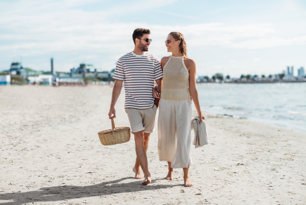 leisure, relationships and people concept - happy couple with picnic basket and blanket walking along beach in tallinn, estonia. happy couple with picnic basket walking on beach