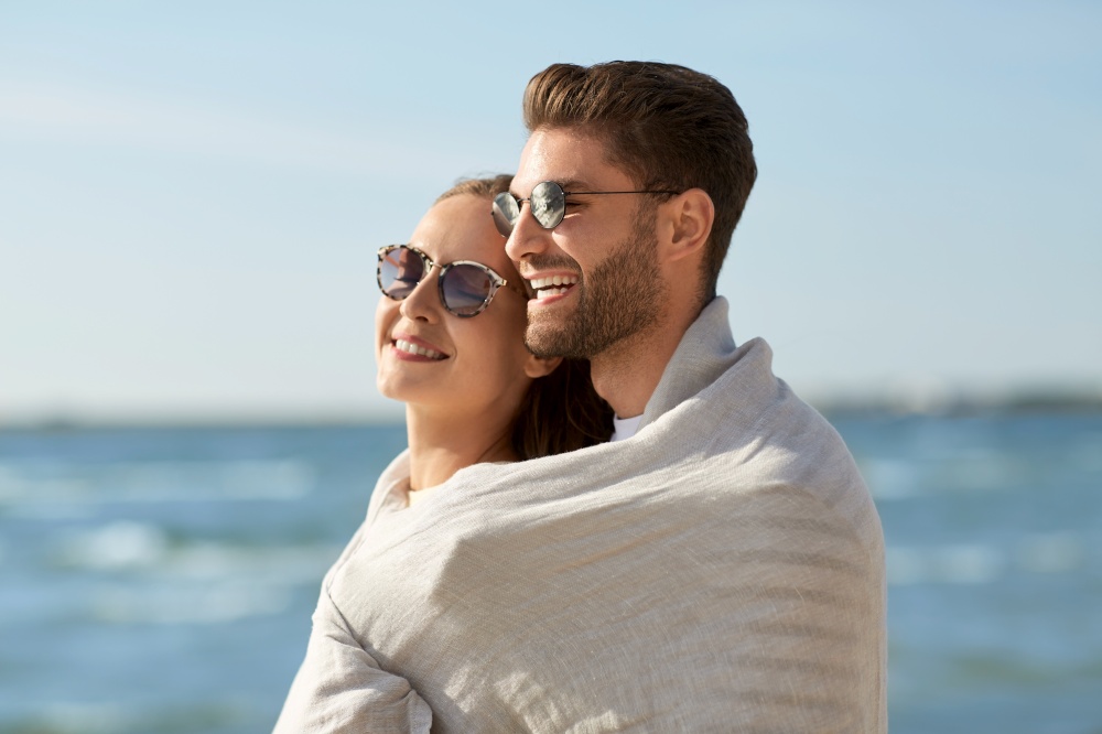 leisure, relationships and people concept - happy couple in sunglasses covered with blanket hugging on summer beach. happy couple covered with blanket hugging on beach