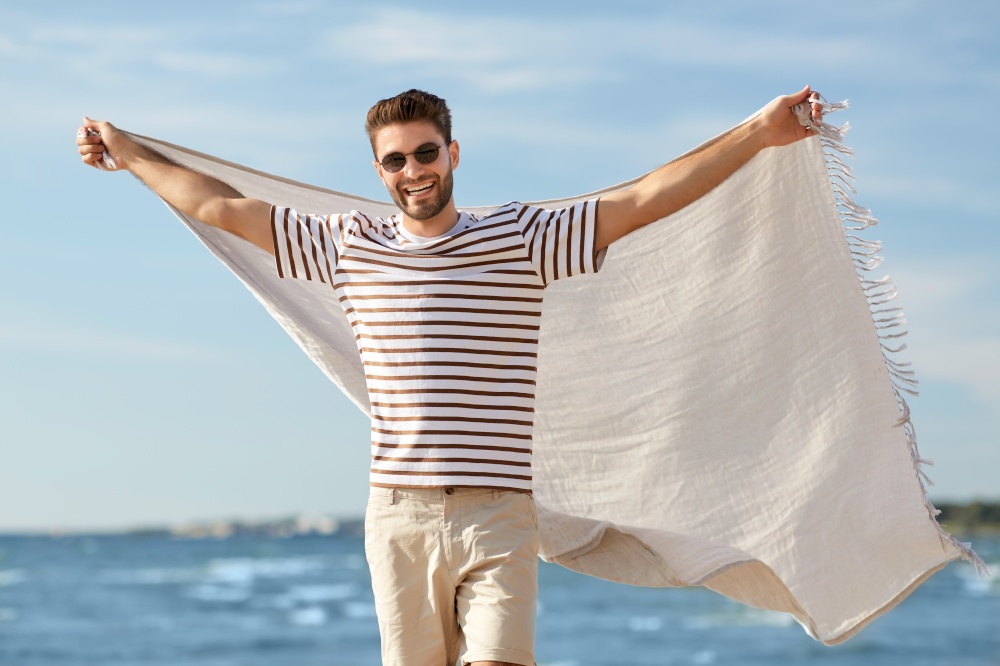 summer holidays and people concept - portrait of happy smiling young man in sunglasses with blanket on beach. smiling man in sunglasses with blanket on beach