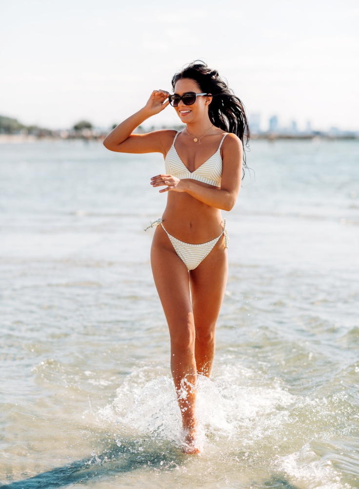 people, summer and swimwear concept - happy smiling young woman in bikini swimsuit running in water beach. smiling young woman in bikini swimsuit on beach
