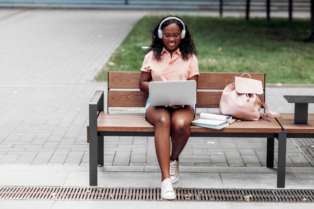 technology, education and people concept - happy smiling african american student girl in headphones with laptop computer and books in city. african student girl in headphones with laptop