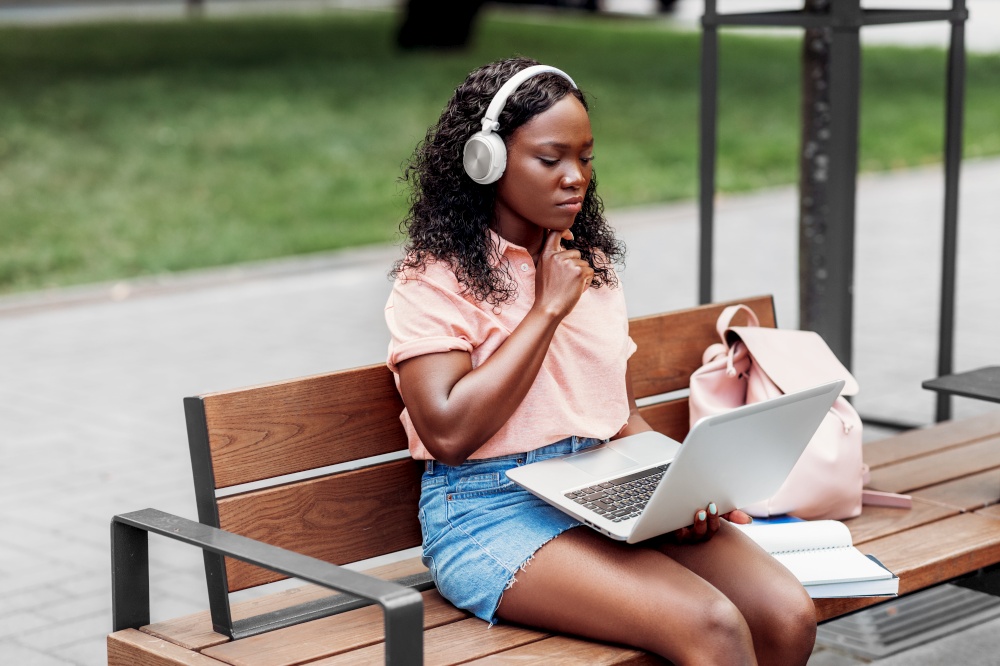 technology, education and people concept - african american student girl in headphones with laptop computer and books in city. african student girl in headphones with laptop