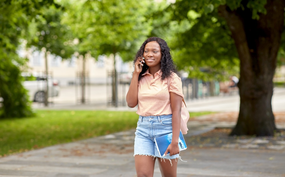 technology, education and people concept - happy smiling african american student girl with textbook calling on smartphone in city. african student girl calling on smartphone in city