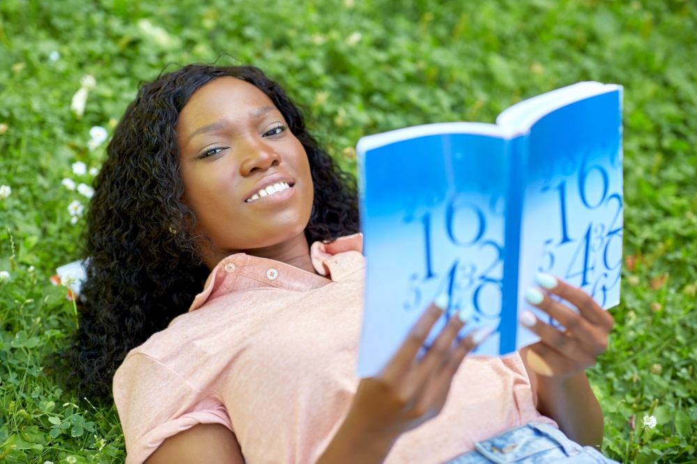 education, school and people concept - happy smiling african american student girl reading math textbook lying on grass. african student girl reading math textbook