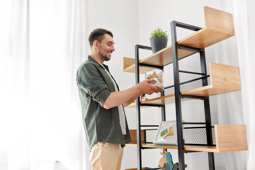 home improvement and decoration and people concept - happy smiling man placing seashells in vase to shelf. man decorating home with seashells in vase