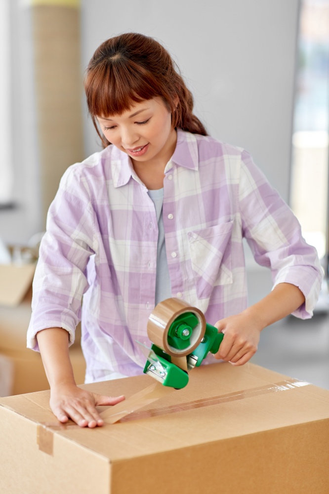 moving, people and real estate concept - happy smiling asian woman with adhesive tape packing box at new home. woman with adhesive tape packing box at new home