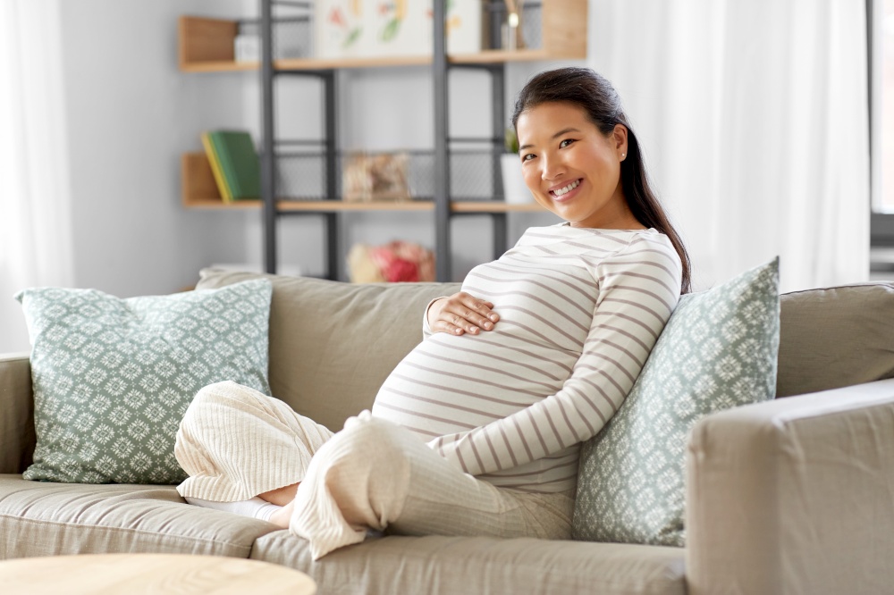pregnancy, rest, people and expectation concept - happy smiling pregnant asian woman sitting on sofa at home. happy pregnant asian woman sitting on sofa at home