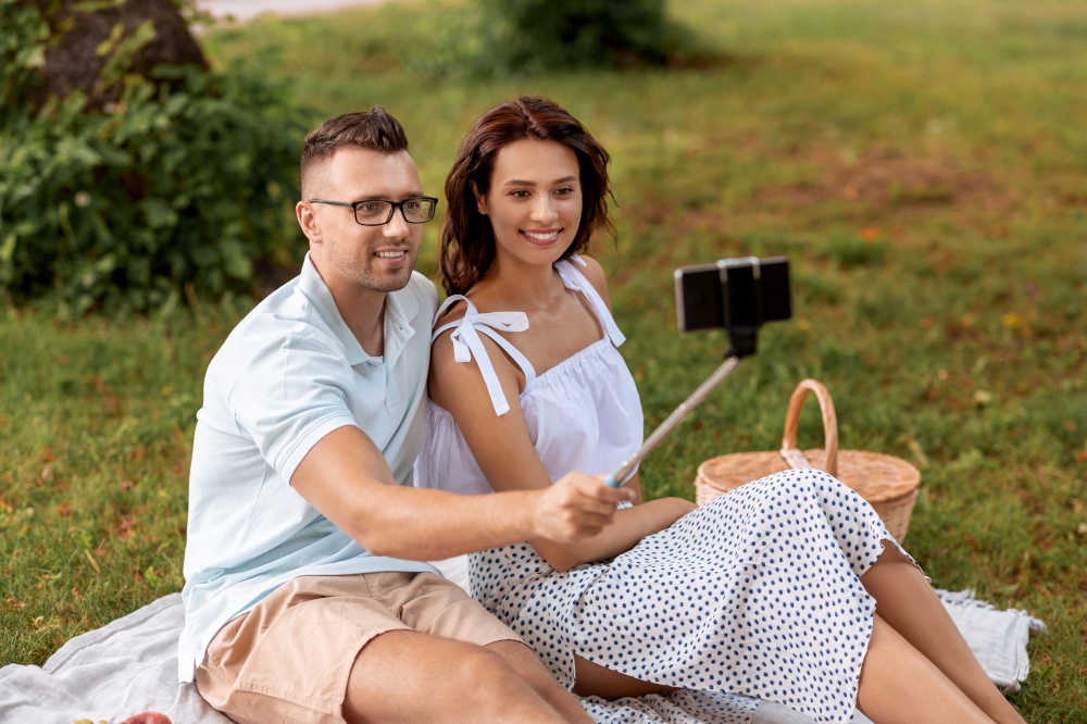 leisure and people concept - happy couple having picnic and taking picture with smartphone on selfie stick at summer park. happy couple taking selfie at picnic in park